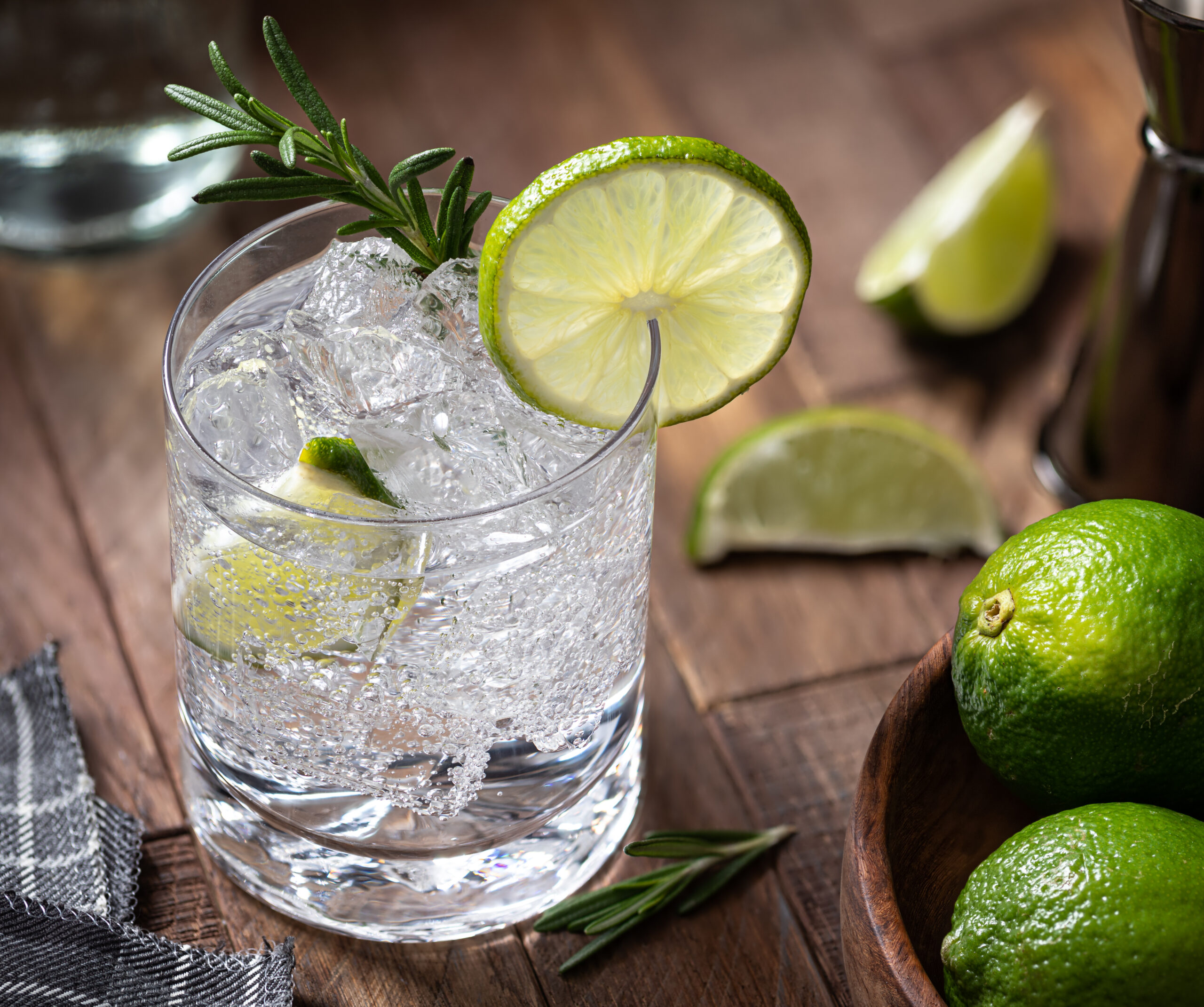 Gin and tonic cocktail with lime slices, rosemary and ice on a rustic wooden table